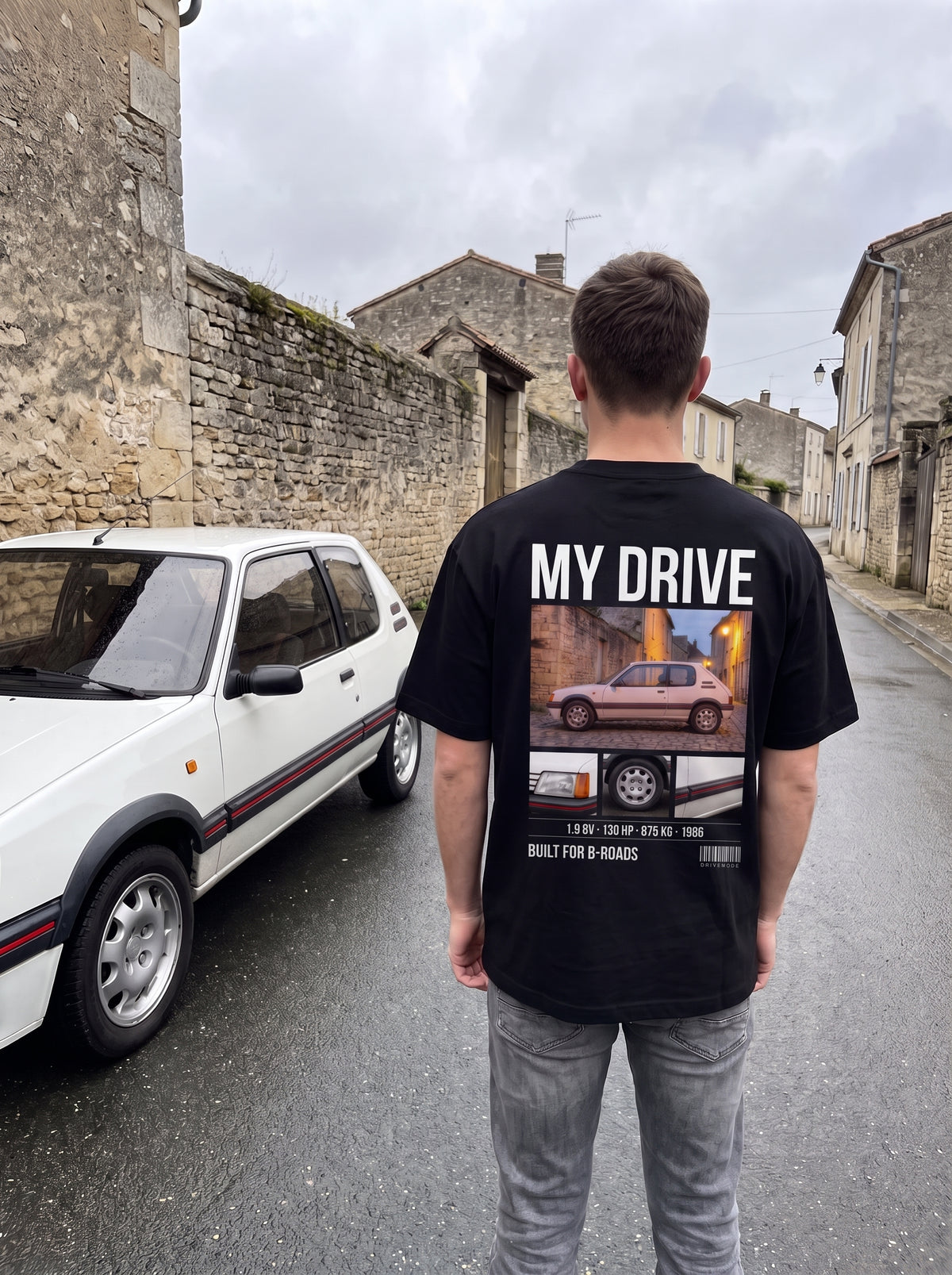 T-shirt personnalisé DriveMode noir oversize porté de dos à côté de la voiture imprimée en collage au dos, dans une rue de village en France