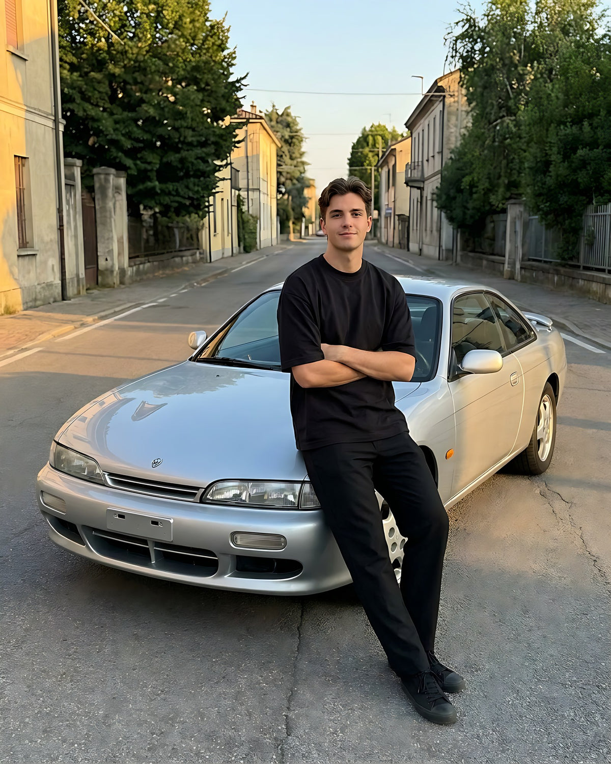 Jeune homme en t-shirt oversize noir DriveMode adossé à un coupé japonais argenté au coucher du soleil