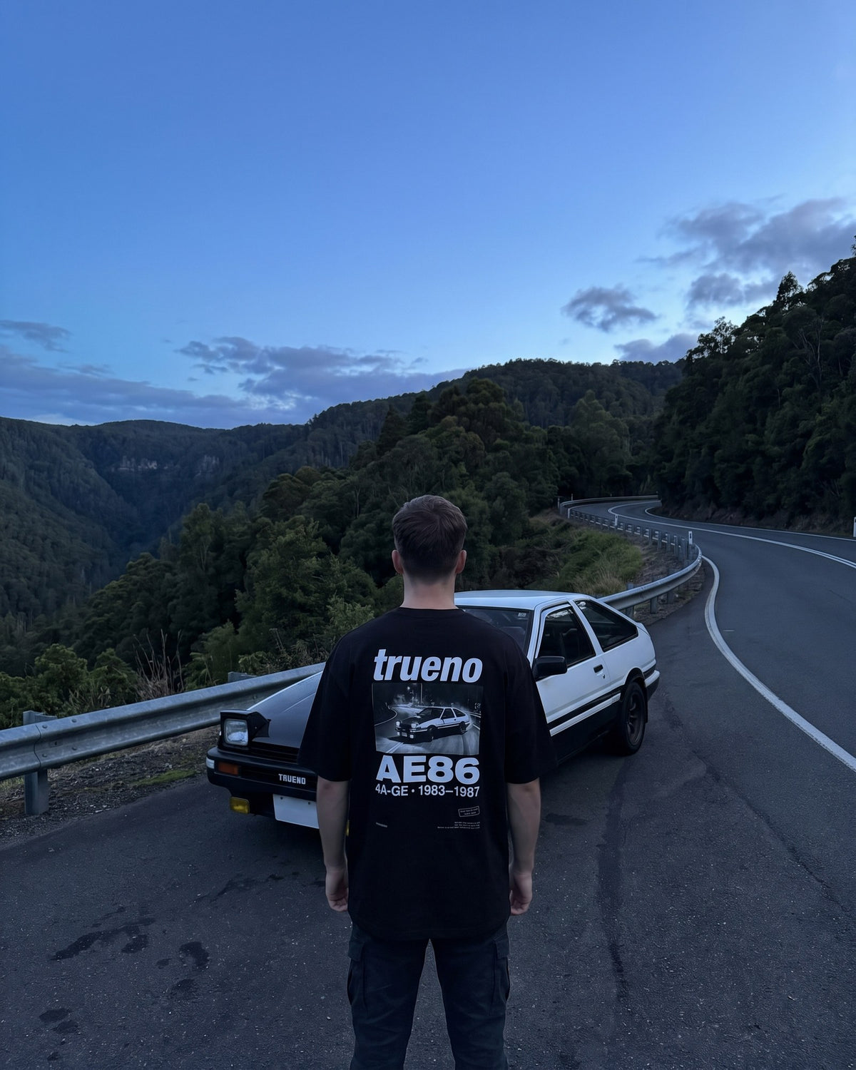 T-shirt oversize Trueno AE86 porté de dos devant une AE86 sur une route de montagne au crépuscule